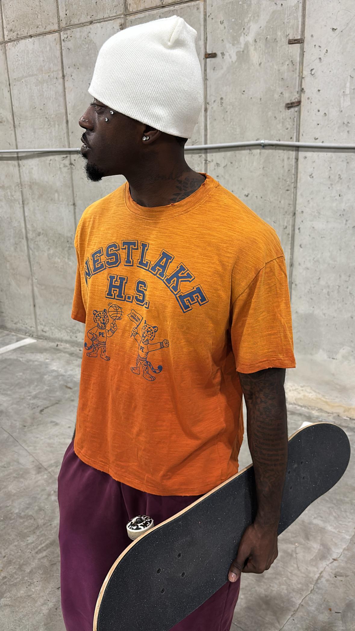 Model in the orange Westlake H.S. Tee holding a skateboard against a concrete wall